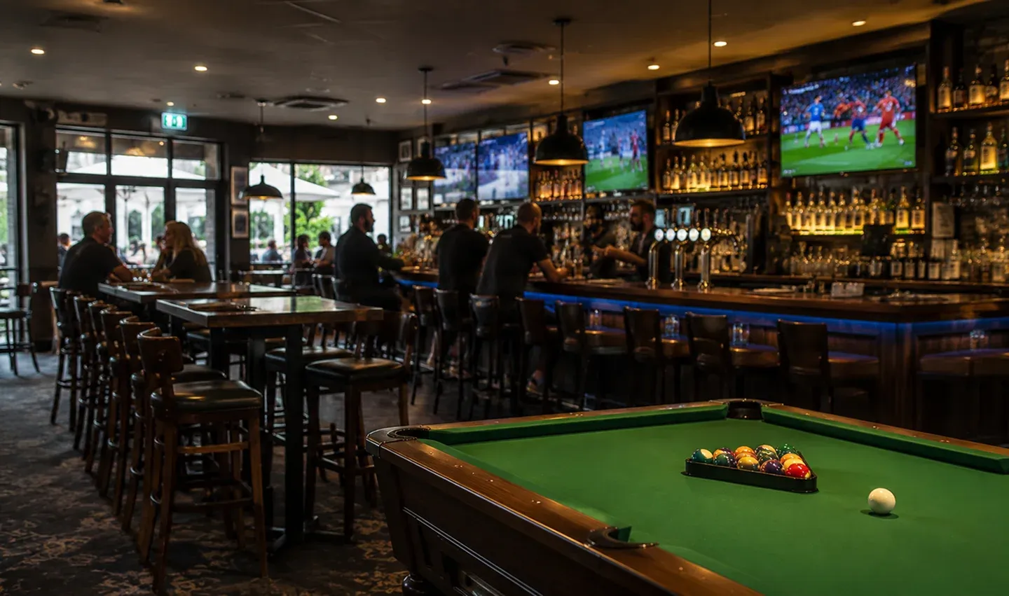 Interior of a dimly lit bar with a pool table in the foreground and patrons sitting at the bar and tables, watching soccer on multiple TVs.